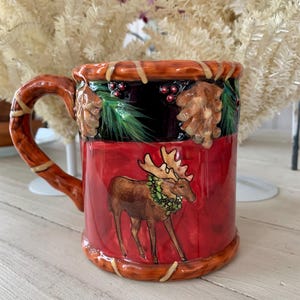 May include: A ceramic mug with a rustic design. The mug features a red base with a brown moose wearing a green wreath. The top rim is black with pinecones, berries, and green pine needles. The handle and rim are brown with a woven design.