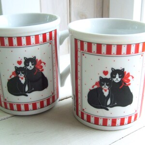 May include: Two white ceramic mugs with red and white stripes. Each mug features a black and white illustration of two cats with red bows and a red heart.