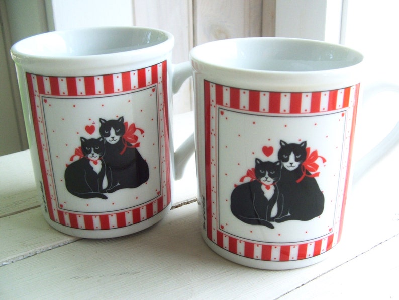 May include: Two white ceramic mugs with red and white stripes. Each mug features a black and white illustration of two cats with red bows and a red heart.