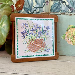 May include: A square ceramic tile featuring a floral design of purple irises in a woven basket, set within a wooden frame. The tile has a white background with a green border. The frame is made of light brown wood.