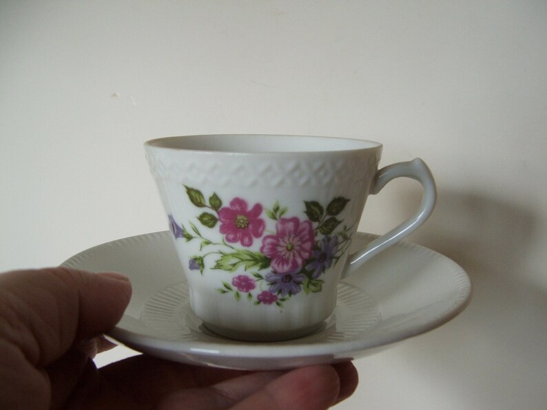 Pode incluir: Uma ch&aacute;vena de ch&aacute; e um pires brancos com um padr&atilde;o floral em tons de rosa, roxo e verde. A ch&aacute;vena tem uma pega delicada e uma borda festonada.