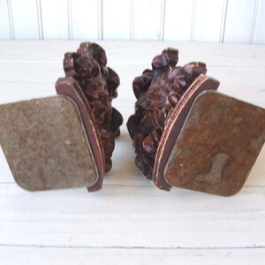 May include: A pair of wooden bookends with intricate carvings of leaves and flowers. The bookends have a dark brown finish and are in the shape of a half-circle. The bookends are sitting on a white surface.