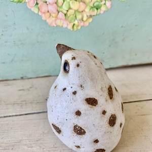May include: A ceramic bird figurine with a white body and brown spots. The bird has a brown beak and a dark eye. The figurine is on a light-colored wooden surface. The background features a floral design.