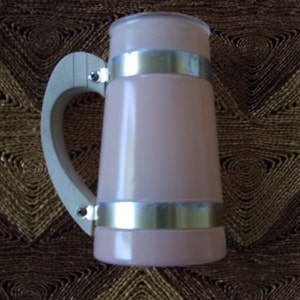 May include: A pink glass beer stein with a wooden handle and silver metal bands. The stein is sitting on a brown woven surface.