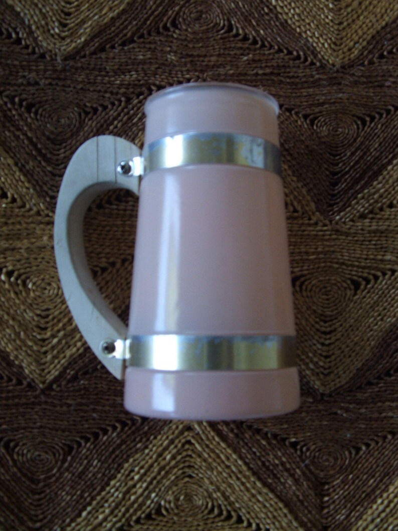 May include: A pink glass beer stein with a wooden handle and silver metal bands. The stein is sitting on a brown woven surface.