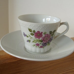 Pode incluir: X&iacute;cara de ch&aacute; e pires de porcelana branca com um padr&atilde;o floral. A x&iacute;cara tem um delicado desenho floral com flores rosa, roxas e verdes. O pires &eacute; branco liso.