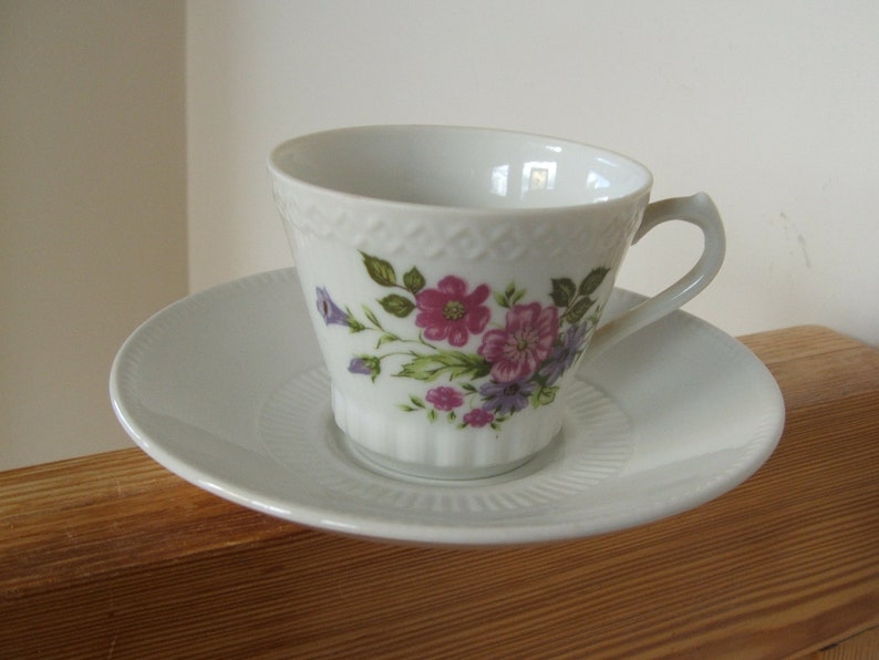 Pode incluir: X&iacute;cara de ch&aacute; e pires de porcelana branca com um padr&atilde;o floral. A x&iacute;cara tem um delicado desenho floral com flores rosa, roxas e verdes. O pires &eacute; branco liso.