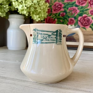 May include: A cream-colored ceramic creamer with a green and brown design. The design features a stylized wooden gate with the text "The Gateway Inn" on it.