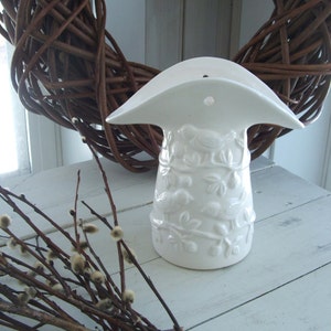 May include: White ceramic wall vase with a bird and floral design. The vase has a hole in the top for hanging. The vase is sitting on a white wooden surface with a branch of pussy willow.