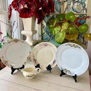 May include: Three antique decorative plates and a small creamer with floral patterns. The plates are displayed on black stands. A white vase with dark red flowers and a green plant are in the background.