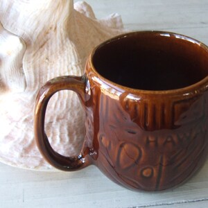 May include: A brown ceramic mug with a raised design of a tiki face and the word "Hawaii".