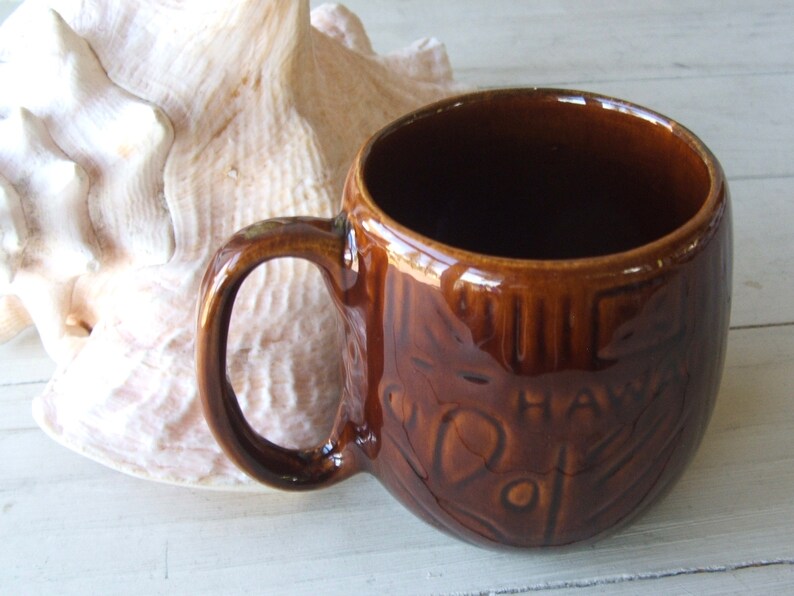 May include: A brown ceramic mug with a raised design of a tiki face and the word "Hawaii".