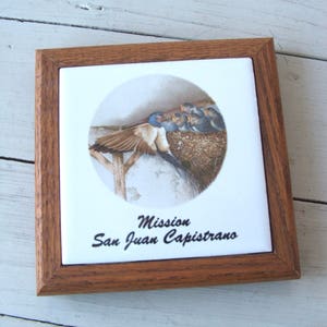 May include: A framed tile with a watercolor painting of a bird family in a nest. The tile is brown with a white background and the text "Mission San Juan Capistrano" is printed below the image.