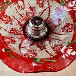 May include: A vintage glass centerpiece bowl with a red rim and floral design. The bowl features a silver metal flower frog in the center. The red rim is decorated with white and pink roses and green leaves. The bowl has a scalloped edge.