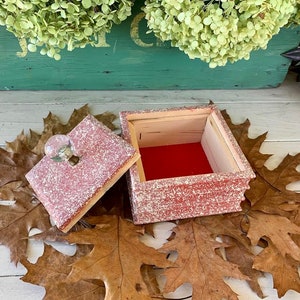 May include: A small, square, red and white speckled wooden box with a clear glass knob on the lid. The box is open and the inside is lined with red felt. The box is sitting on a bed of brown leaves.
