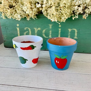 May include: Two painted terracotta flower pots, one white with green and red peppers, the other blue with a red strawberry. The pots are sitting on a white wooden surface in front of a green wooden box with the word "jardin" painted on it.