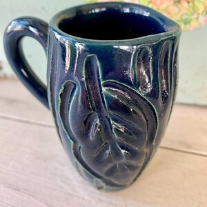 May include: A handmade ceramic mug in a deep blue glaze with a leaf design. The mug has a curved handle and a textured surface. The interior is dark blue, and the exterior has hints of green.