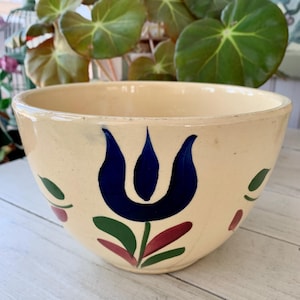 May include: Cream-colored ceramic bowl with a hand-painted floral design. The bowl features a large blue tulip with green leaves and red accents. The bowl is sitting on a white wooden surface.