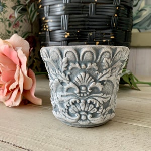May include: A small, gray ceramic planter with an ornate, embossed floral design. The planter has a slightly flared rim and a rounded base.