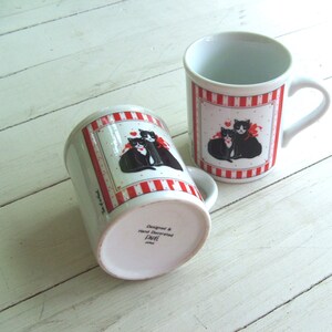 May include: Two white ceramic mugs with red and white stripes. Each mug features a black and white illustration of two cats with a red heart. The bottom of one mug reads "Designed & Hand Decorated, Laurel, Japan".