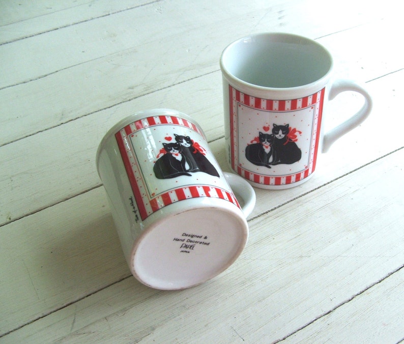 May include: Two white ceramic mugs with red and white stripes. Each mug features a black and white illustration of two cats with a red heart. The bottom of one mug reads "Designed & Hand Decorated, Laurel, Japan".