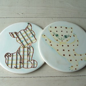 May include: Two ceramic coasters with animal illustrations. One coaster features a brown and white dog with a plaid pattern. The other coaster features a white cat with brown spots.