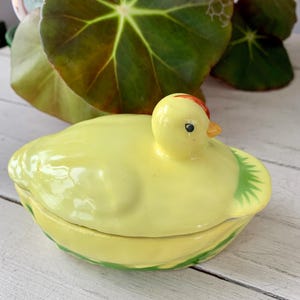 May include: A yellow ceramic duck-shaped trinket box with a lid. The duck has a red crest and an orange beak. The box is decorated with green accents and sits on a white wooden surface. The overall theme is a decorative home item.