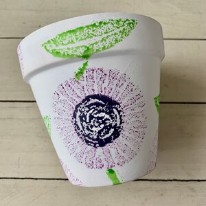 May include: White terracotta flower pot with a purple and green floral design. The design is created with a stamp or stencil.