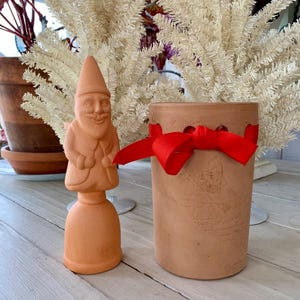 May include: A terracotta gnome figurine stands next to a cylindrical terracotta pot tied with a red ribbon. The gnome has a pointed hat and a smiling face. The pot is empty. The background includes a white decorative tree and a terracotta pot.