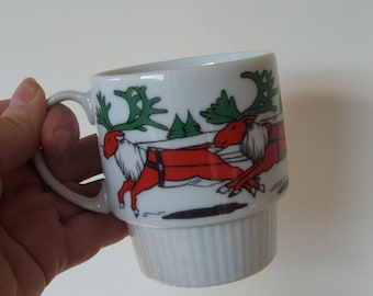 May include: White ceramic mug with a red, black, and green reindeer design. The reindeer are pulling a sled with a red and black design.