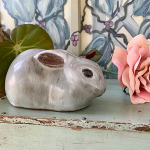 May include: A ceramic rabbit figurine in shades of gray with brown accents, positioned next to a pink rose. The bunny has detailed features and sits on a distressed, light blue surface. Decorative home decor.