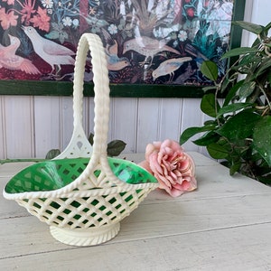 May include: A white ceramic basket with a green liner and a woven handle. The basket is empty and sitting on a white wooden table.