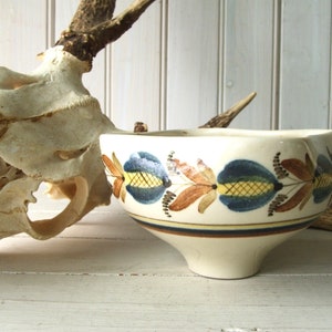 May include: A white ceramic bowl with a blue, yellow, and brown floral design. The bowl has a wide rim and a narrow base.