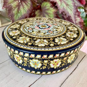 May include: A round, navy blue tin with a floral design. The lid and sides feature white and yellow flowers with gold accents. The center of the lid has a light blue and pink floral pattern.