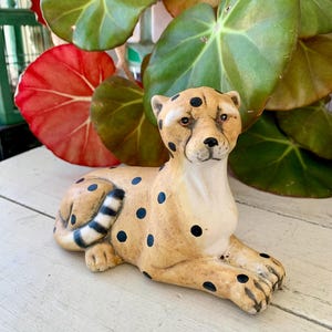May include: A ceramic cheetah figurine in a resting pose, featuring a tan body with black spots and a striped tail. The figurine is set against a backdrop of green and red plant leaves, creating a naturalistic setting.