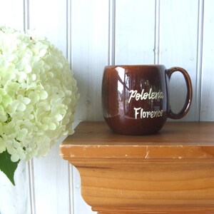 May include: A brown ceramic mug with a white inscription that reads "Pololena Florence". The mug is sitting on a wooden shelf.