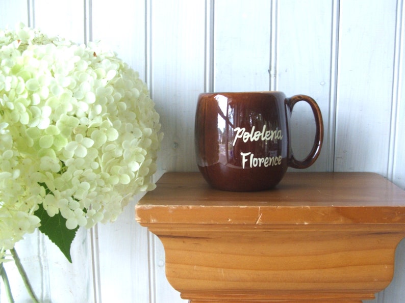 May include: A brown ceramic mug with a white inscription that reads "Pololena Florence". The mug is sitting on a wooden shelf.