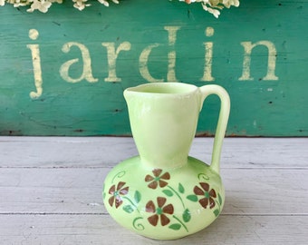 May include: A small, light green ceramic pitcher with a brown floral design. The pitcher has a rounded body and a small spout. The word "jardin" is written on a green wooden surface behind the pitcher.