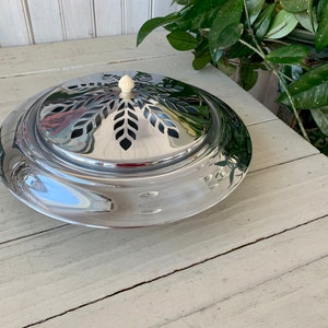 May include: A silver, round, decorative dish with a leaf pattern and a white knob on top. The dish is sitting on a white wooden surface.