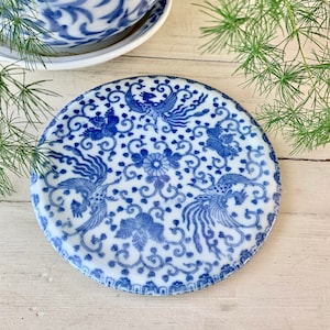 May include: A blue and white ceramic coaster with a floral and bird design. The coaster is round and has a raised edge.