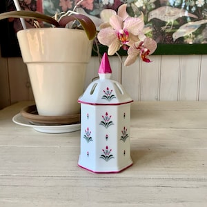 May include: A white, hexagonal ceramic candle holder with a pink top and trim. The candle holder is decorated with a repeating floral pattern in pink and gray. The background includes a potted orchid and a floral print.