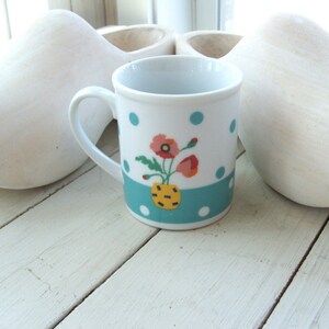 Puede incluir: Una taza de cer&aacute;mica blanca con un patr&oacute;n de lunares azules y blancos y una banda turquesa. La taza tiene un dise&ntilde;o floral con amapolas rojas, amarillas y blancas en un jarr&oacute;n amarillo.