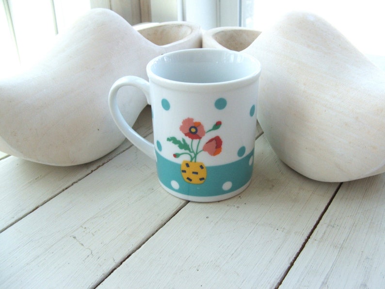 Puede incluir: Una taza de cer&aacute;mica blanca con un patr&oacute;n de lunares azules y blancos y una banda turquesa. La taza tiene un dise&ntilde;o floral con amapolas rojas, amarillas y blancas en un jarr&oacute;n amarillo.