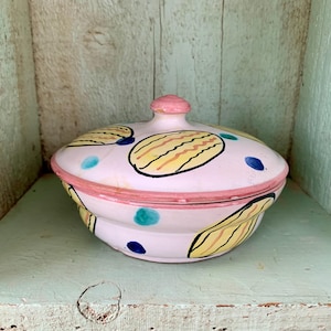 May include: A ceramic trinket dish with a lid, painted in pastel pink with yellow and black oval designs, teal and navy blue dots, and a pink rim. The lid has a small pink knob.