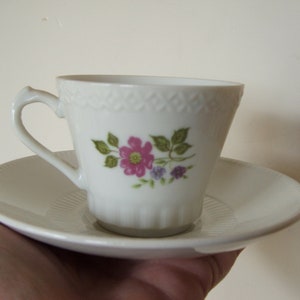 Pode incluir: Um conjunto de ch&aacute;vena e pires de porcelana branca com um design floral com flores rosa e roxas e folhas verdes.