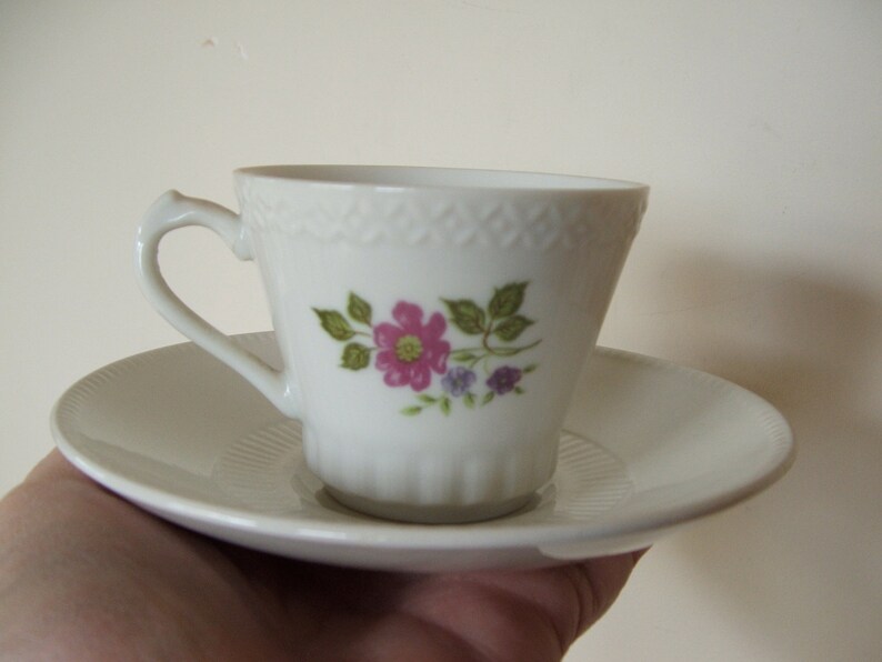 Pode incluir: Um conjunto de ch&aacute;vena e pires de porcelana branca com um design floral com flores rosa e roxas e folhas verdes.