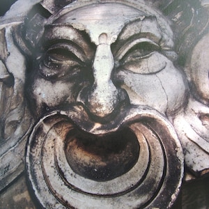 May include: A close-up of a weathered stone sculpture of a face with a large, open mouth. The sculpture is carved with intricate details, including a prominent nose and deep-set eyes. The mouth is framed by a circular, ring-like structure.