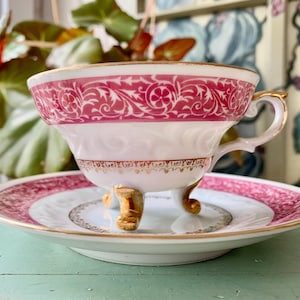 May include: A vintage teacup and saucer set. The white teacup features a pink floral design and gold trim. The saucer has a matching pink border and gold accents. The teacup is elevated by three gold feet.