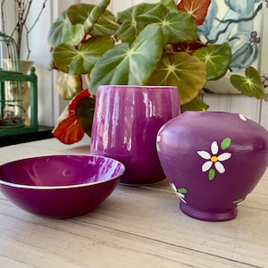 May include: A collection of decorative purple ceramic items. Includes a bowl with a white rim, a cylindrical vase, and a small vase decorated with white and yellow flower designs. The items are arranged on a light-colored wooden surface.