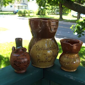 May include: Three brown ceramic jugs with painted designs. The largest jug has the text "Restaurante la Trucha Madrid" on it. The smallest jug has the text "Estuve en Algeciras" on it. The middle jug has the text "Robanie con sabor de familia" on it.